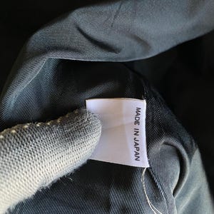 May include: Close-up of a white tag with the text "MADE IN JAPAN" in black, attached to a dark fabric. The fabric appears to be a dark, possibly black, material with a subtle texture. The tag is rectangular and has a clean, simple design.