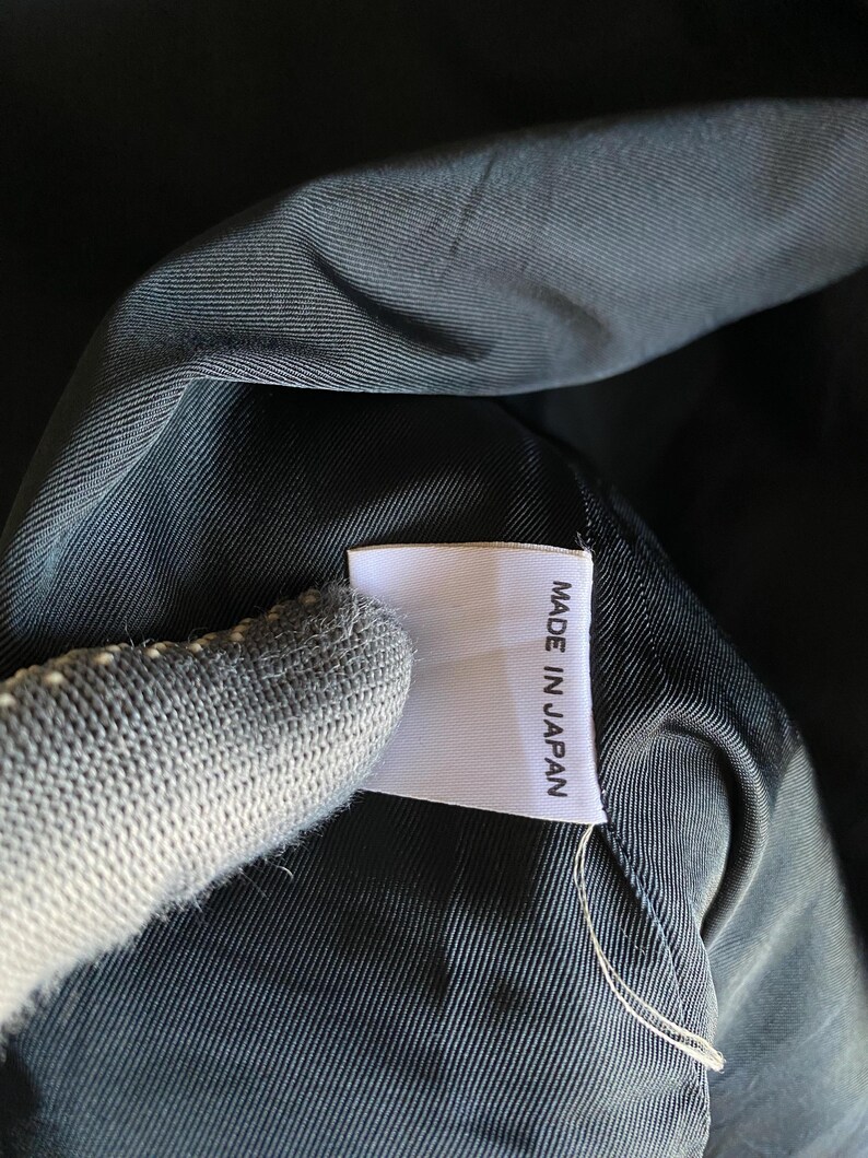 May include: Close-up of a white tag with the text "MADE IN JAPAN" in black, attached to a dark fabric. The fabric appears to be a dark, possibly black, material with a subtle texture. The tag is rectangular and has a clean, simple design.