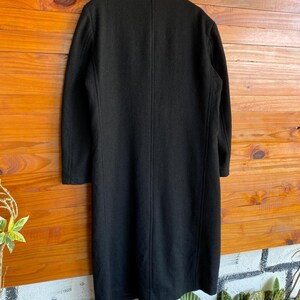 May include: A long, black coat hanging on a wooden surface. The coat has a classic design with a collar and long sleeves. The coat appears to be made of a wool or wool-blend fabric. The coat is a solid black color.