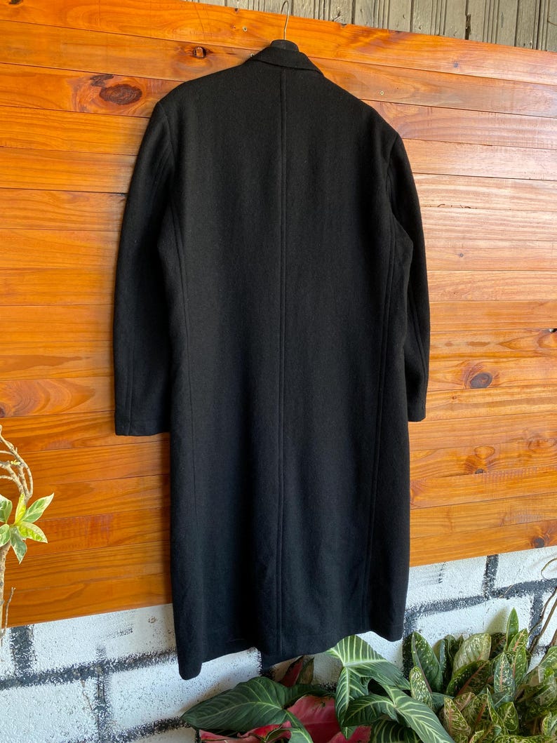 May include: A long, black coat hanging on a wooden surface. The coat has a classic design with a collar and long sleeves. The coat appears to be made of a wool or wool-blend fabric. The coat is a solid black color.