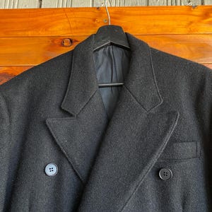 May include: A black double-breasted coat with two rows of buttons and a notched lapel. The coat is hanging on a black hanger against a wooden background. The coat has a classic, tailored design.