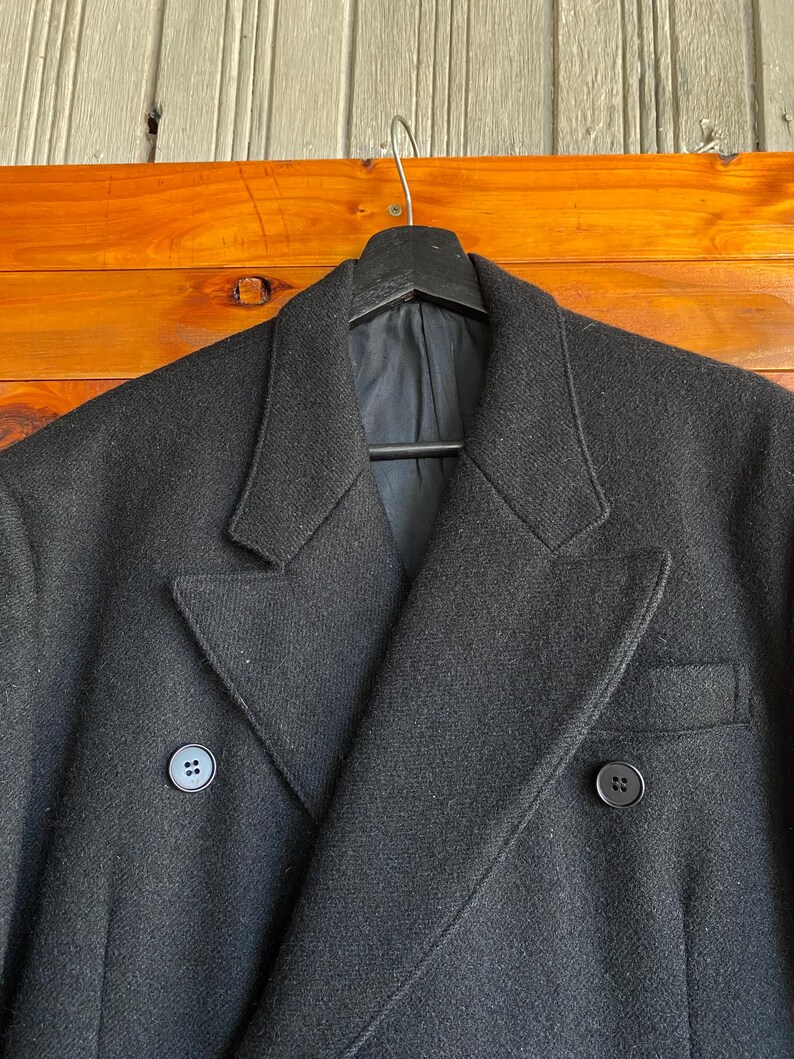 May include: A black double-breasted coat with two rows of buttons and a notched lapel. The coat is hanging on a black hanger against a wooden background. The coat has a classic, tailored design.