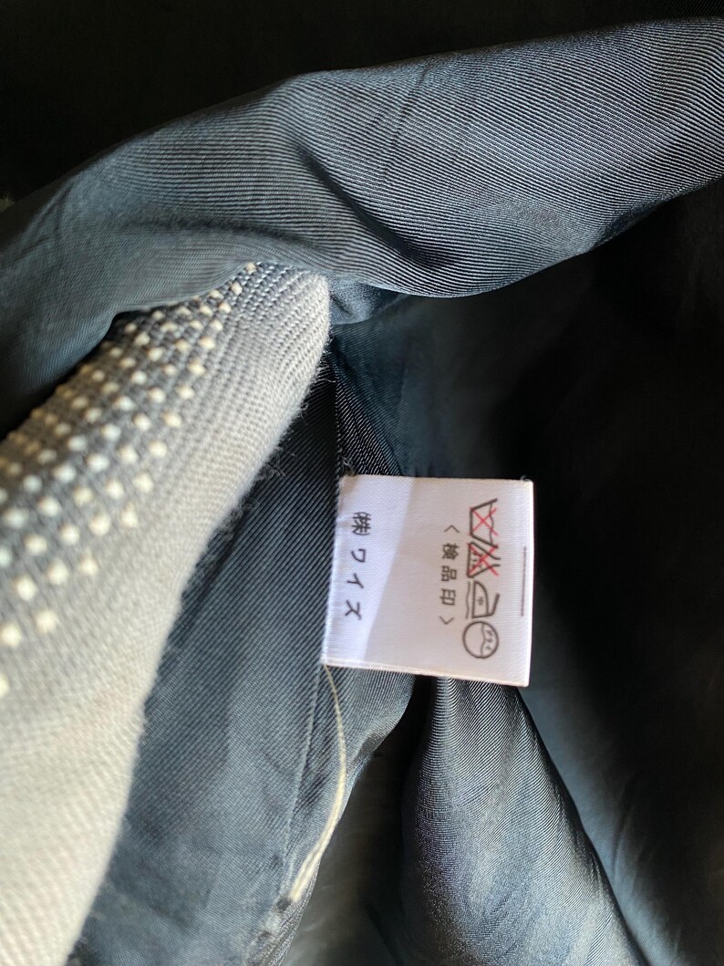 May include: Close-up of a garment's interior, showcasing a label with Japanese text and care symbols. The fabric appears to be a dark, possibly navy, color. The label is white with black text and symbols.
