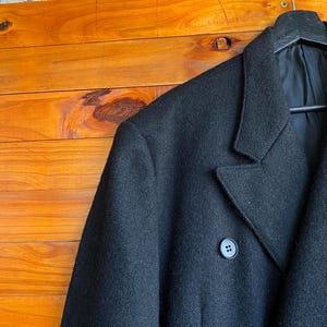 May include: A black wool coat with a notched lapel and a two-button closure. The coat is hanging on a black hanger against a wooden background. The coat's fabric appears to be a solid, dark color.