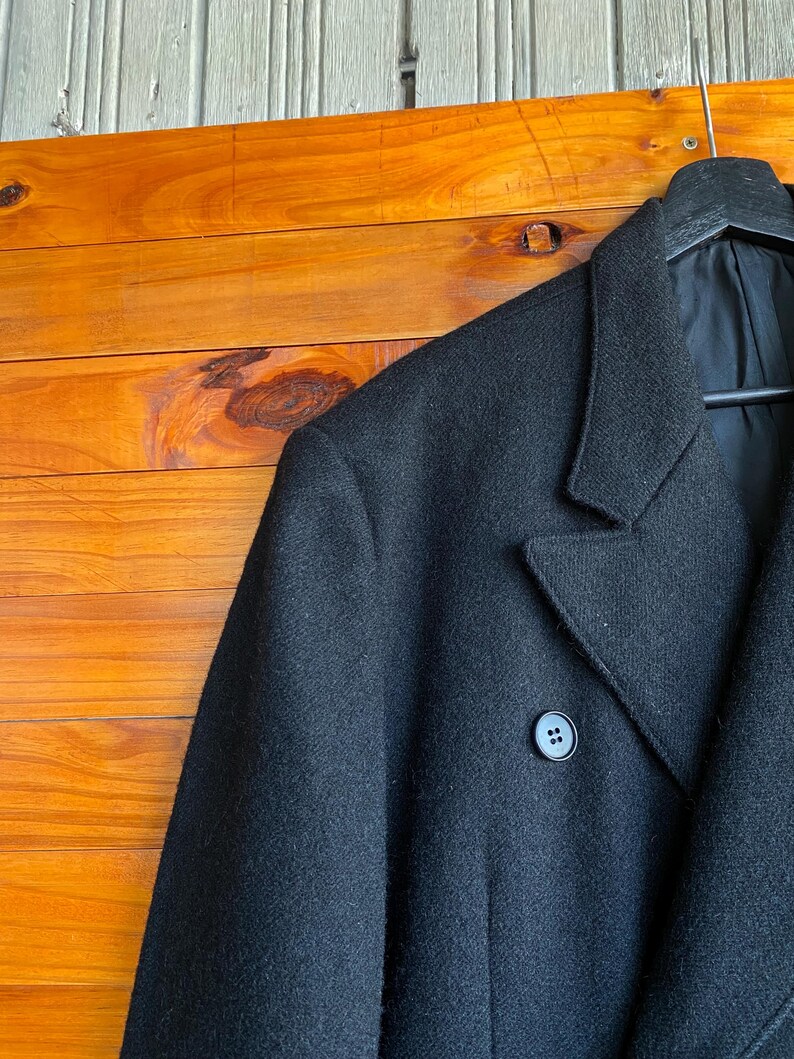 May include: A black wool coat with a notched lapel and a two-button closure. The coat is hanging on a black hanger against a wooden background. The coat's fabric appears to be a solid, dark color.
