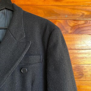 May include: Close-up of a black wool blazer with a two-button closure and a notched lapel. The blazer has a single chest pocket and a dark blue lining. The garment is hanging on a black hanger against a wooden background.