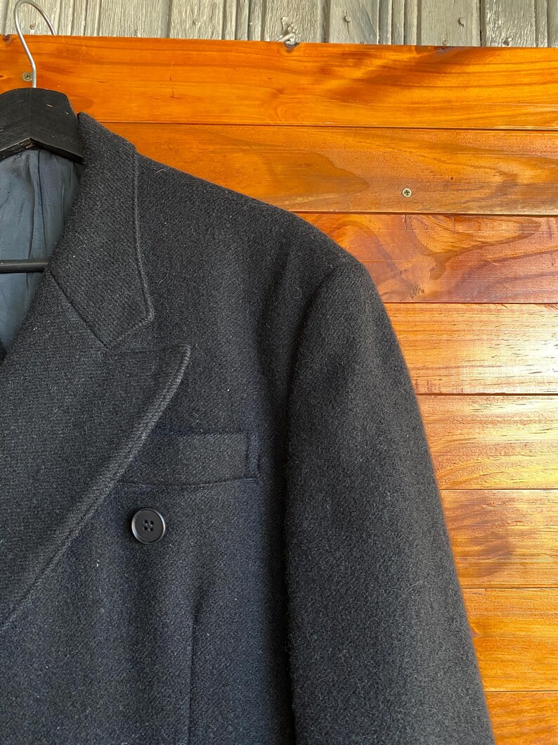 May include: Close-up of a black wool blazer with a two-button closure and a notched lapel. The blazer has a single chest pocket and a dark blue lining. The garment is hanging on a black hanger against a wooden background.