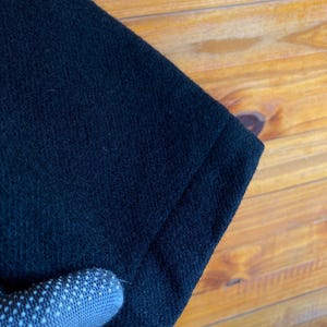 May include: Close-up of a section of black fabric, possibly a garment. The fabric has a textured surface and appears to be a dark, solid color. The edge of the fabric is visible, showing a folded seam. The background is a wooden surface.