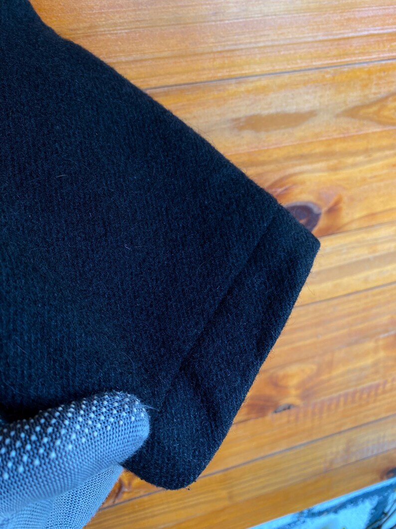 May include: Close-up of a section of black fabric, possibly a garment. The fabric has a textured surface and appears to be a dark, solid color. The edge of the fabric is visible, showing a folded seam. The background is a wooden surface.