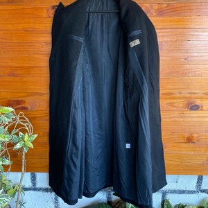 May include: A black wool coat hanging on a black hanger. The coat is open, revealing a black lining and interior pockets. The coat is displayed against a wooden background.