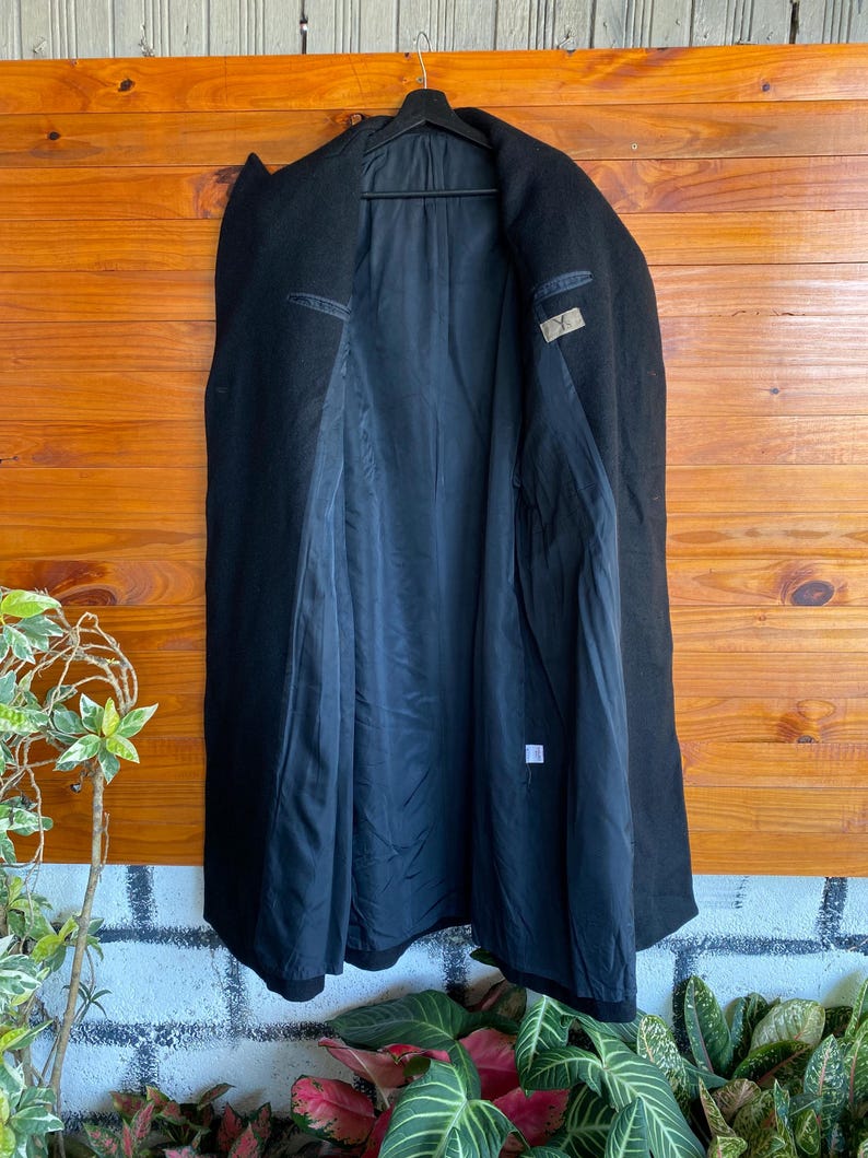 May include: A black wool coat hanging on a black hanger. The coat is open, revealing a black lining and interior pockets. The coat is displayed against a wooden background.