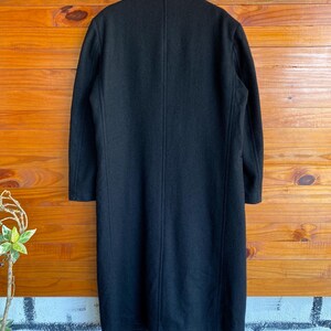 May include: A long, black wool coat with a classic design. The coat features a notched collar, long sleeves, and a straight silhouette. The back of the coat has a center vent for ease of movement. The coat is displayed against a wooden background.
