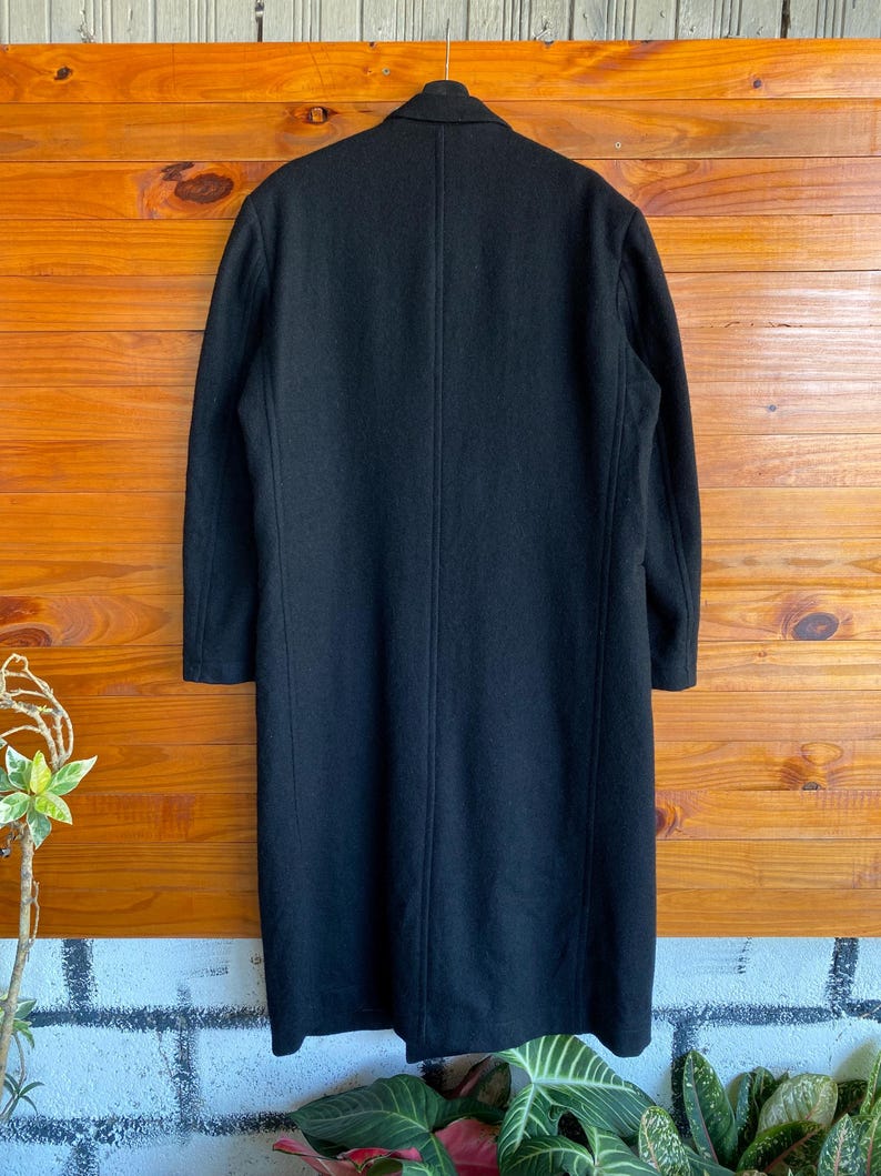 May include: A long, black wool coat with a classic design. The coat features a notched collar, long sleeves, and a straight silhouette. The back of the coat has a center vent for ease of movement. The coat is displayed against a wooden background.