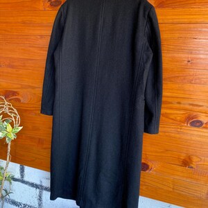 May include: A long, black wool coat hangs against a wooden background. The coat has a classic design with a notched collar and a single-breasted closure. The coat's length extends to the mid-calf, and it has a tailored fit.