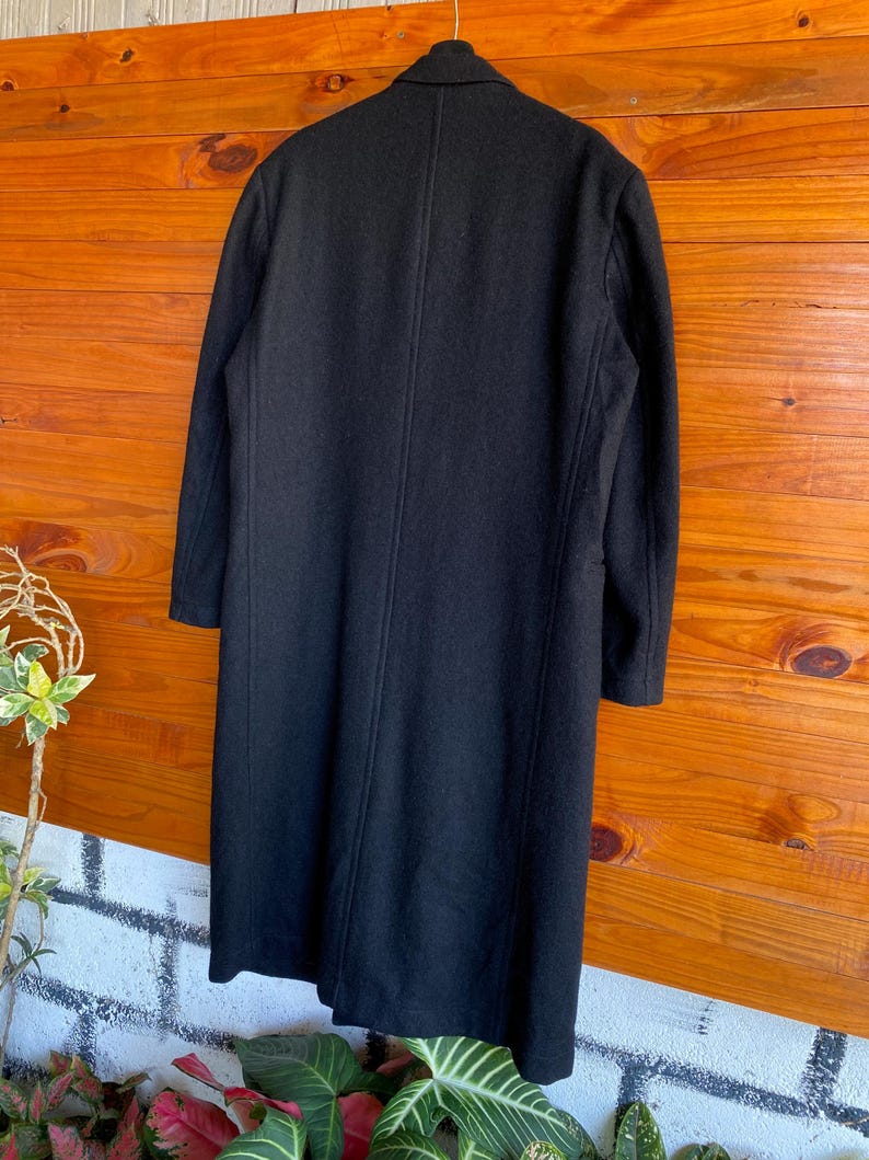 May include: A long, black wool coat hangs against a wooden background. The coat has a classic design with a notched collar and a single-breasted closure. The coat's length extends to the mid-calf, and it has a tailored fit.