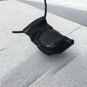 Op de afbeelding: Een zwart leren tasje met een gevlochten bandje dat aan een wit oppervlak hangt.