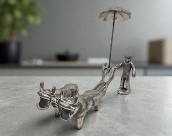 Peut inclure: Figurine argentée représentant un homme tenant un parapluie, tirant deux bœufs. Les bœufs sont attelés ensemble. La figurine est détaillée et semble être en métal.