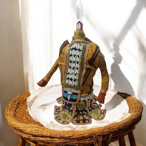 Può includere: Una bambola fatta a mano che indossa un costume tradizionale con intricati dettagli di perline e conchiglie. La bambola è vestita con un tessuto marrone e giallo con una gonna adornata con paillettes e perline. La bambola è in piedi su una base intrecciata.