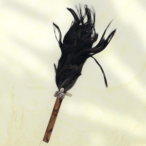 May include: A decorative wand featuring a cluster of black feathers atop a wooden handle. The handle is brown with a light-colored band near the feathers. A small, white shell is attached to the base of the feathers.