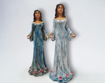 Könnte beinhalten: Zwei Keramikfiguren von Frauen in langen Kleidern. Die Kleider sind in Blau- und Weißtönen bemalt, mit roten Rosen am Saum. Jede Figur trägt eine Krone und hält einen Edelstein in jeder Hand. Die Figuren sind dekorativ und detailliert.