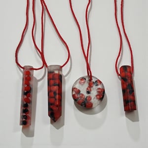 Puede incluir: Cuatro collares de resina hechos a mano con cordones rojos. Los colgantes son de resina transparente con cuentas rojas y negras. Un colgante es redondo, dos son rectangulares y uno tiene forma de cristal.