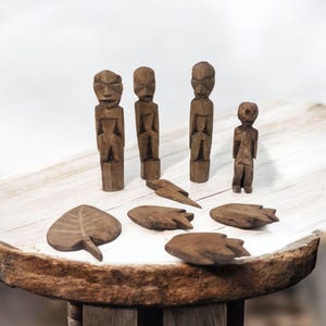 Könnte beinhalten: Ein Set aus fünf Holzfiguren, darunter vier große Figuren mit geschnitzten Gesichtern und eine kleinere Figur. Die Figuren sind auf einem Holztisch angeordnet, zusammen mit fünf weiteren Holzstücken in Form von Blättern und einem Blitz.