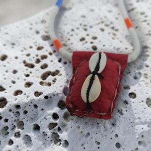 Pode incluir: Um colar com um pendente em forma de bolsa vermelha texturizada, adornado com duas conchas de búzios brancas costuradas verticalmente com fio preto. O cordão é branco com detalhes em azul e laranja, contrastando com o material vermelho.