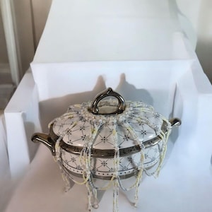Pode incluir: Um prato de servir de cerâmica branca com uma borda dourada e um design de contas. O prato tem uma tampa com uma alça dourada e é decorado com contas brancas e douradas.