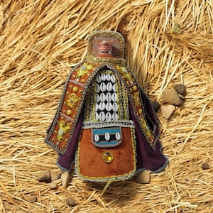 Può includere: Una bottiglia decorativa con una copertura in tessuto marrone, viola e oro. La bottiglia è decorata con conchiglie bianche e bordi dorati. La bottiglia è appoggiata su un letto di paglia.