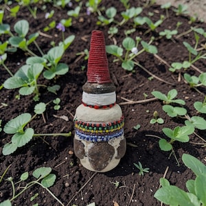 May include: A decorative bottle stopper with a distinctive design. It has a red, conical top, a white midsection, and a base decorated with colourful beads. The base is a light tan colour with brown accents.