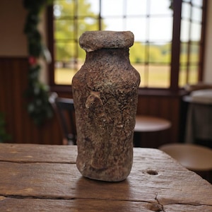 May include: A weathered, brown ceramic vase with a textured surface. The vase has a wide, rounded top and a narrow neck. It sits on a rustic wooden table. The background shows a window and outdoor scenery.