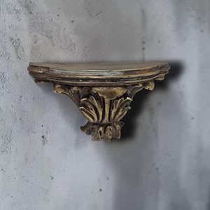 以下が含まれることがあります： 金色の仕上げが施された、小さく装飾的な壁棚。 棚は前面が湾曲しており、葉のような装飾的なブラケットで支えられています。 棚は灰色の壁に取り付けられています。