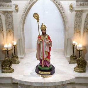 Può includere: Una statuetta di Sant'Agostino, un santo cattolico, in piedi su una base verde con un'iscrizione dorata che recita "Sto. Agostinho". La statuetta indossa una veste rossa e rosa e tiene un bastone sormontato da una croce. La statuetta è davanti a un arco in marmo bianco con due candelabri dorati su entrambi i lati.