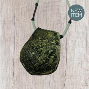 Op de afbeelding: Een unieke ketting met een getextureerde hanger in groen- en bruintinten. De hanger hangt aan een wit koord met zwarte en bruine accenten. Op de achtergrond staat een turquoise cirkel met de tekst "NEW ITEM".