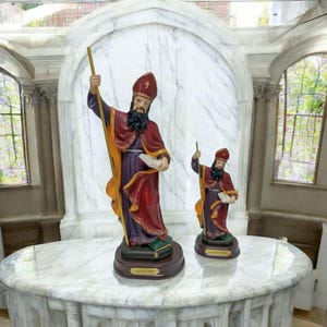 Può includere: Due statue religiose di Sant'Agostino, una più grande dell'altra, entrambe in piedi su una superficie di marmo bianco. La statua più grande tiene un libro e un bastone, mentre la statua più piccola tiene un libro e un bastone. Entrambe le statue indossano una veste rossa e viola con bordi dorati. Le statue si trovano in un ambiente di chiesa con una parete di marmo bianco e una finestra di vetro colorato sullo sfondo.