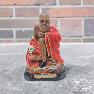 Könnte beinhalten: Eine bemalte Figur einer kahlen Person mit roten Augen, die ein leuchtend rotes Gewand mit Goldbesatz trägt. Die Figur sitzt auf einem Sockel, ein kleiner Totenkopf liegt auf dem Schoß. Eine rechteckige Plakette ist am schwarzen Sockel befestigt.