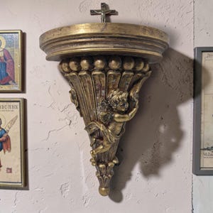 以下が含まれることがあります： 金色の装飾的な壁棚。 天使のデザインが特徴です。 棚は丸い上部と装飾的な溝付きサポートを備えています。 小さな十字架が棚の上に設置されています。 テクスチャ加工された薄いピンク色の壁に取り付けられています。