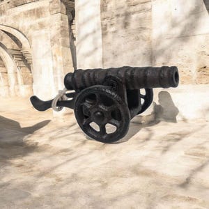 May include: A black cast iron cannon with wheels, a barrel, and a handle. The cannon is sitting on a stone surface.