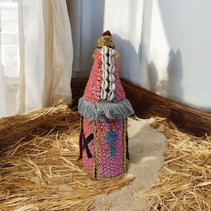 Può includere: Un oggetto decorativo a forma di bottiglia rosa e oro ricoperto di paillettes e conchiglie. L'oggetto ha una frangia grigia intorno alla parte superiore e una croce nera sul davanti.