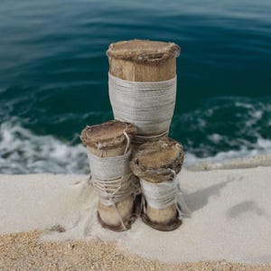 May include: Three wooden drums with white string wrapped around them. The drums are sitting on a white sandy beach with blue water in the background.