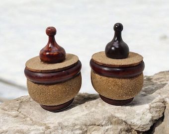 May include: Two small wooden containers with cork lids and wooden finials. The containers are brown and have a sandy texture. They are sitting on a light gray rock.