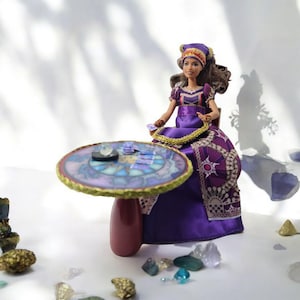 May include: A purple and gold doll dressed as a fortune teller sits at a table with a crystal ball and tarot cards. The table has a stained glass design and a burgundy base. The doll has brown hair and is wearing a purple and gold head scarf.