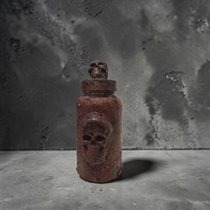 Könnte beinhalten: Eine zylindrische braune Flasche mit einer texturierten, verwitterten Oberfläche. Die Flasche zeigt einen markanten geprägten Totenkopf auf der Vorderseite und eine kleinere Totenkopffigur auf dem Deckel, was ein einheitliches, dunkles Aussehen ergibt.