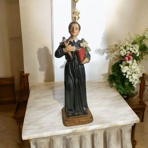 Könnte beinhalten: Eine weiße Statue eines jungen Jungen in einer schwarzen Robe, der ein Buch und ein Lamm hält. Die Statue steht auf einem weißen Marmoraltar mit einem Blumenschmuck dahinter.