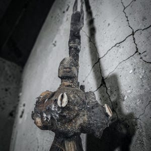 Puede incluir: Una escultura artesanal oscura y texturizada que presenta una cabeza y un torso de aspecto humano. La superficie está incrustada con materiales orgánicos y adornada con tres conchas de cauri blancas. Una borla oscura y fibrosa cuelga de la base.