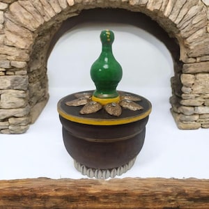 May include: A small, round, brown wooden box with a green and yellow lid. The lid has a decorative top with a green finial and gold accents. The box is decorated with gold leaf and has a white base.