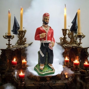 May include: A red and black figurine of a man wearing a red turban and holding a small object in his right hand. The figurine is standing on a green base and is surrounded by candles and smoke.