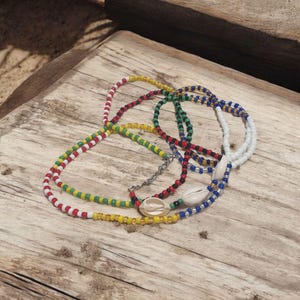Op de afbeelding: Een verzameling kralenkettingen in verschillende kleuren, waaronder rood, geel, groen en blauw, met witte schelpaccenten. De kettingen zijn gerangschikt op een verweerd houten oppervlak, wat een bohemien of strandachtige uitstraling creëert.