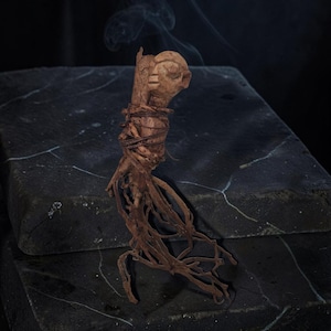 Könnte beinhalten: Eine braune, geschnitzte Holzfigur mit einem abgerundeten Kopf und einer strukturierten, wurzelartigen Basis. Die Figur ist mit dünnem, dunkelbraunem Garn umwickelt und stößt oben einen leichten Rauchschleier aus, was ihr ein organisches Aussehen verleiht.