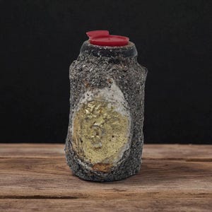 Peut inclure: Une bouteille en céramique grise texturée avec un couvercle rouge. La bouteille présente un motif ovale doré sur le devant. La bouteille est présentée sur une surface en bois sur fond noir.